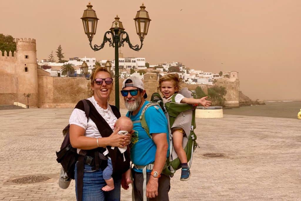 A family are stood with the walls and white medina of Rabat behind them. The mum has on e ahite top and is carrying her baby in a carrier on her front.  The dad is stood next to her with a blue t-shirt on and has their son on his back in a carrier.  They are wearing sunglasses as there is adust storm happening. Behind the family between them and the medina is a lamp post. 