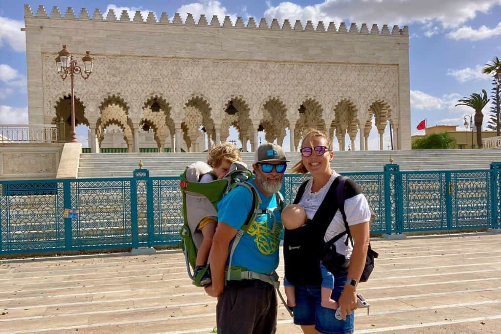 A family is standing outside the Mohammed V Mausoleum.  The mum is on the right with a white top and carrying her baby in a baby carrier.  The man is wearing a blue t-shire, hat and sunglasses and a backpack carrier with his son in it.  The Mausoleum is one of the best things to do in Rabat as it's the resting place of the leader of Morocco.  The building itself is white with intricate carving.