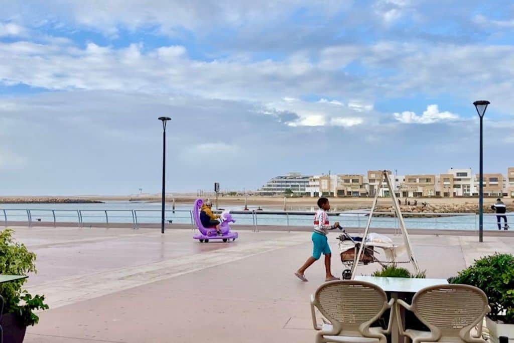 This is at the waterfront near the Medina in Rabat.  There is a man in a purple electric car with his son.  Riding in these cars is one of the fun things to do in Rabat.  In the foreground of the image is a table and chairs as the photo is taken from a restaurant. A local woman is crossing the photo pushing a pram, and in the background is the river and the view over to the city of Sale with it's white buildings.