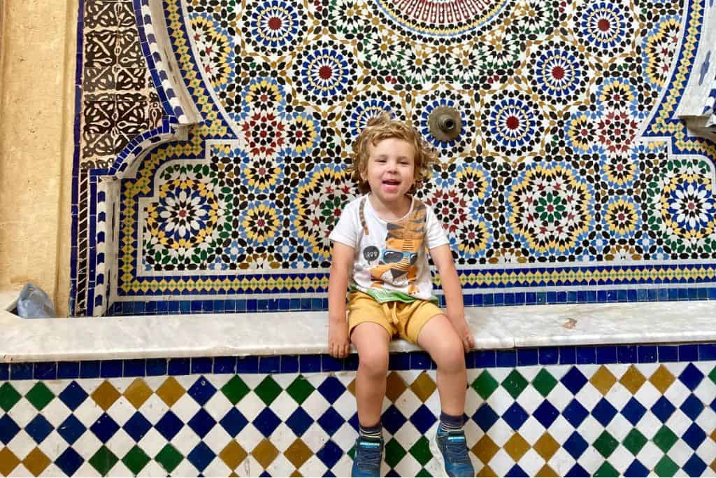 A little bpy in a white t-shirt and yellow shorts is sat on the wall of a water fountain in the kasbah in Rabat in Morocco. 