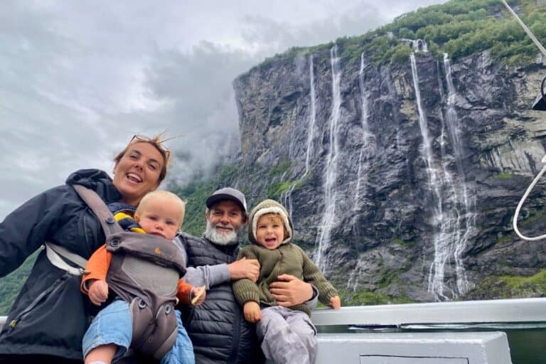 A mum wearing her baby on her in a baby carrier is stood next to her husband who is holding their 3 year old son. They are on the car ferry having a photo take in front of a waterfall in Geirgangerfjord which is one of the best things to do in Geiranger.