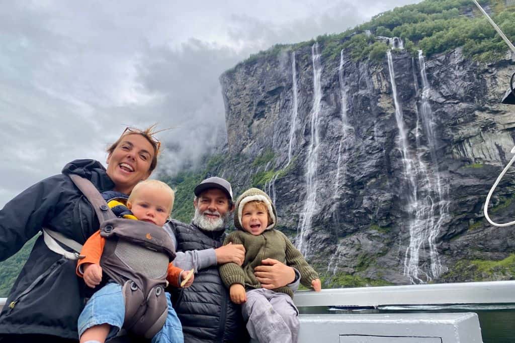 A mum wearing her baby on her in a baby carrier is stood next to her husband who is holding their 3 year old son. They are on the car ferry having a photo take in front of a waterfall in Geirgangerfjord which is one of the best things to do in Geiranger. 