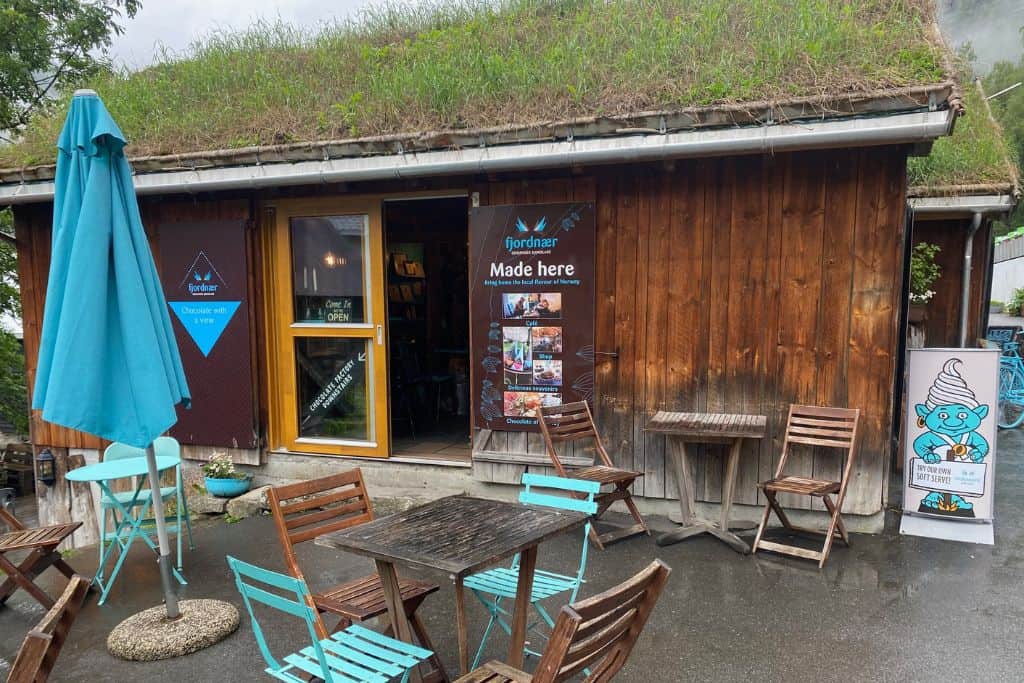 The outside of a brown boat house in Geiranger that has now been turned into a cafe serving chocolate. There is a blu umbrella and some metal garden furniture outside that people can use to eat and drink at the cafe. This is in Geiranger.