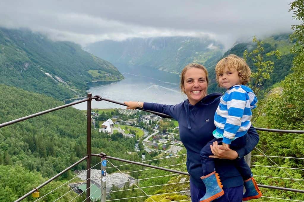 A mum is smiling at the camera and holding her three year old son in her arms.  She's holding onto a metal railing and behind her is the Geirangerfjord in Norway. They are stood at the Flydalsjuvet viewpoint.