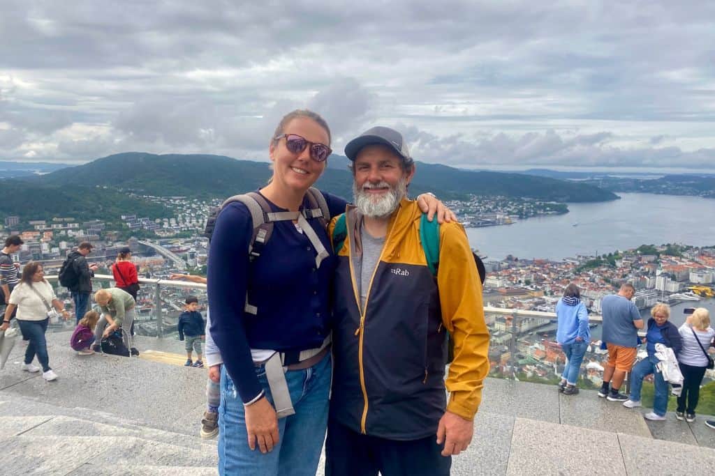 A woman in blue cardigan, jeans and sunglasses has her arm around her boyfriend who has a raincoat on and baseball cap.  They're both smiling at the camera and behind there is the view out over Bergen.  This is the view from the top of Mount Floyen which is one thing that everyone visits if they're in Bergen for one day.