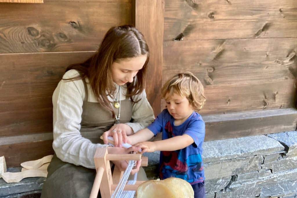 A small boy in a blue t-shirt is helping a girl dressed as a Viking to weave a belt.  She is watching him and he is concentrating.