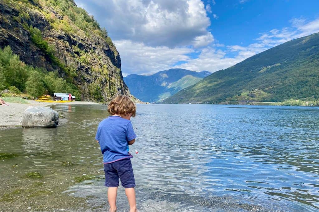 A little boy in blue t-shirt and shorts is standing with his back to the camera with his feet in the water at a pebble beach.  In the background of the image is the fjord in Flam as he is stood on the beach there.