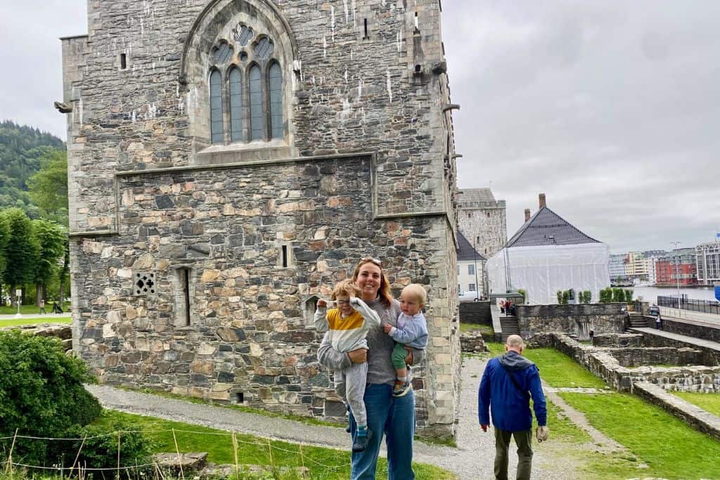 A woman is stood smiling at the camera holding her 1 year old and her 3 year old sons in her arms. Behind her is the fortress in Bergen which is made of stones from 1500s.