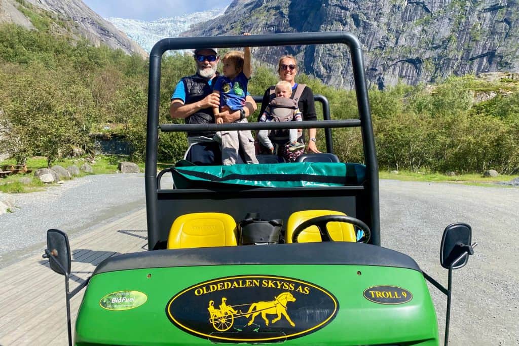 The image is a photo of a large green off road goldf buggy.  In the back is a couple holding their baby and their toddler.  Behind them is the Briksdal glacier which is one of the best things to do in Olden in Norway.