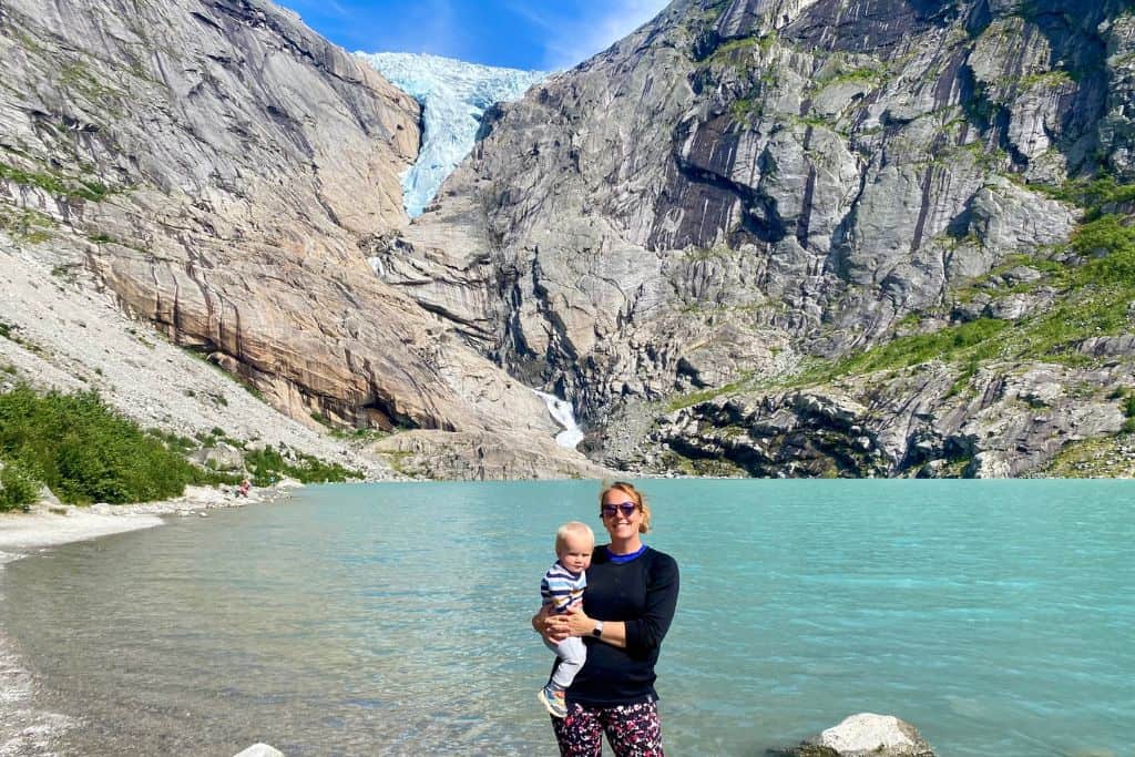 A woman in black top and leggings is holder her baby in her arms and smiling at the camera. She's stood in a lake and behind her is a mountain with the Briksdal glacier coming down into the lake. This is one of the best things to do in Olden in Norway.