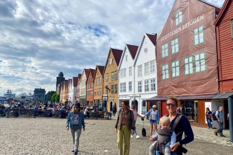 A woman in sunglasses is smiling at the camera whilst wearing her baby in a carrier. She is stood outside the front of the colourful wooden buildings in Bryggen. Visiting Bryggen is a great start to one day in Bergen.