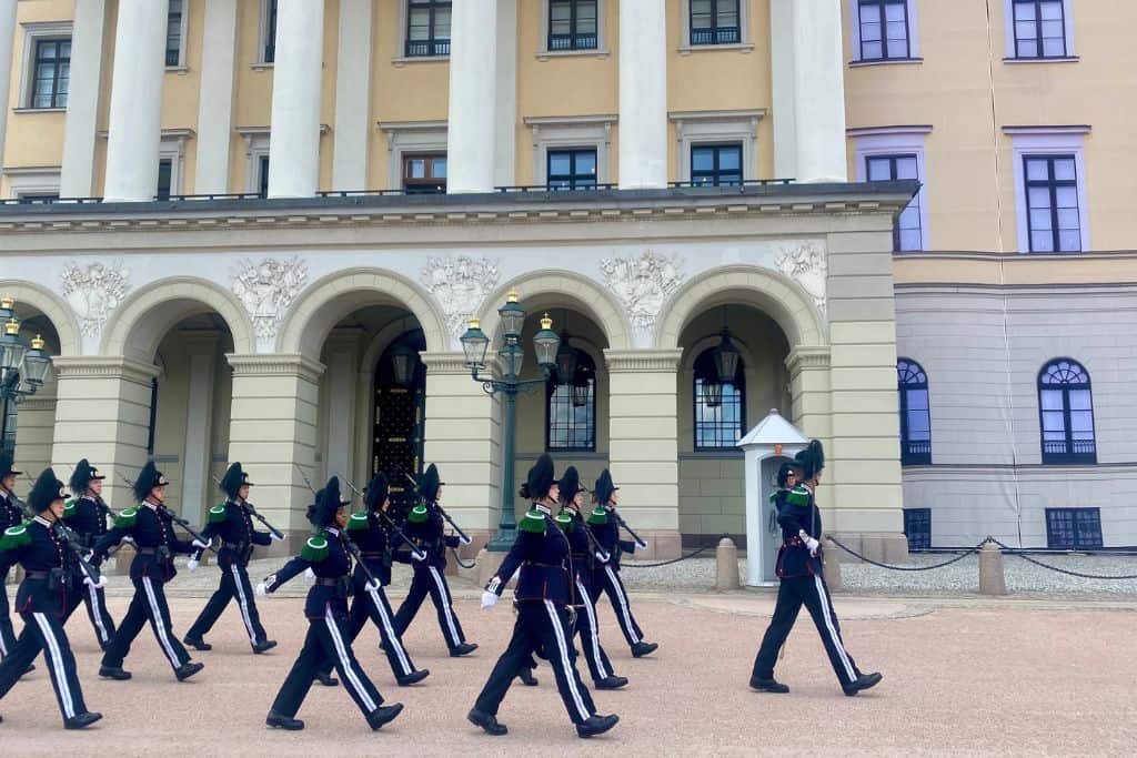 The outside of the Royal Palace in Oslo. In front of the palace the changing of the guards is happening and rows of guards are walking from the left of the image over to the right.