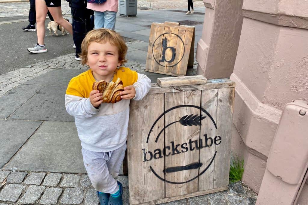 A little boy in a tracksuit i smiling at the camera as he eats a cinnamon bun. He is spending one day in Bergen and this is his breakfast.