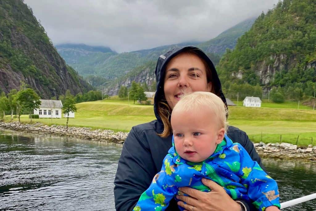 A woman has her black raincoat on with her hood up as it's slightly rainy. In her arms is her son in a dinosaur raincoat.  They're both looking at the camera and behind them is a fjord in Bergen in Norway.