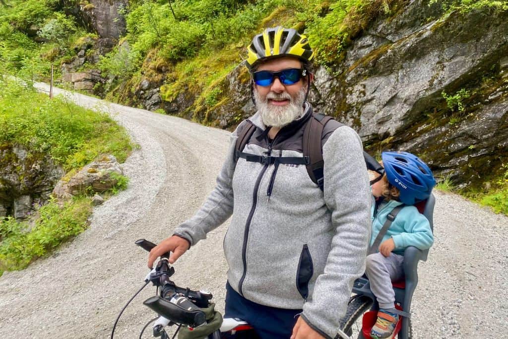 A man with a beard i a grey fleece is looking at the camera with is cycle helmet on.  His is stood astride his bike. One the back of his bike is his 3 year old son.  They are on a track going down a mountain.