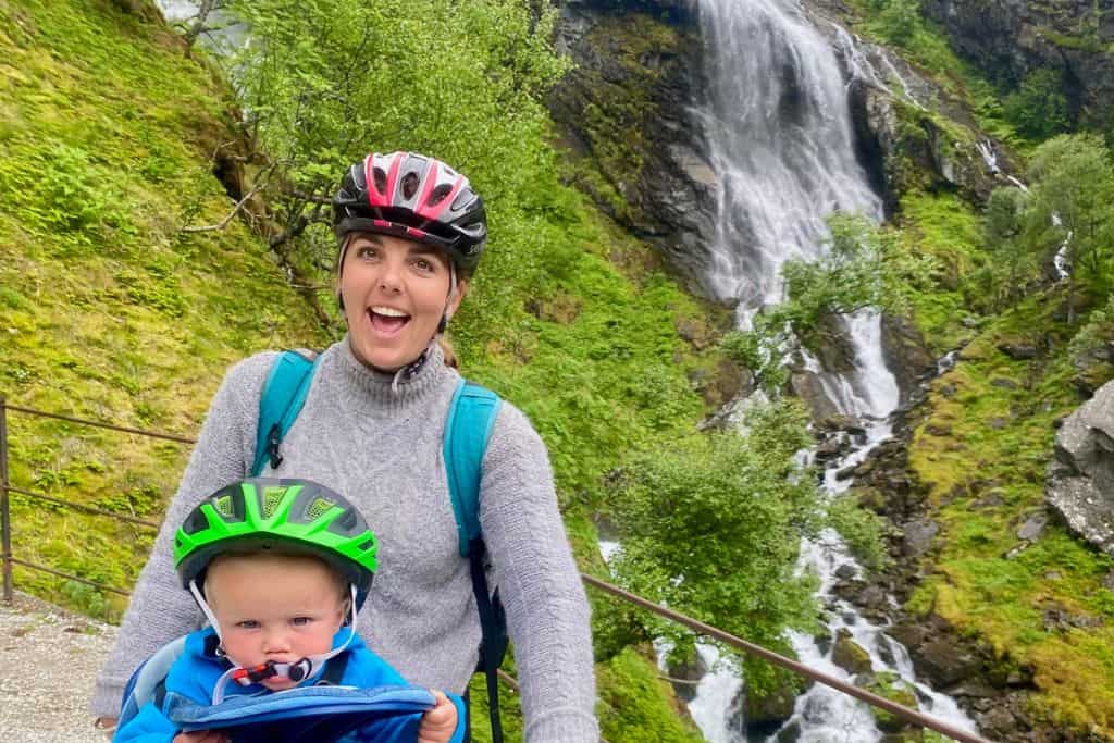 A woman is smiling at the camera in a grey jumper with her cycle helmet. She is standing over her bike and her 1 year old son is sat on the bike in is blue fleece with his bike helmet on.  Behind them is a waterfall.  This is the bike ride down from the Flam railway and is one of the popular things to do in the area.