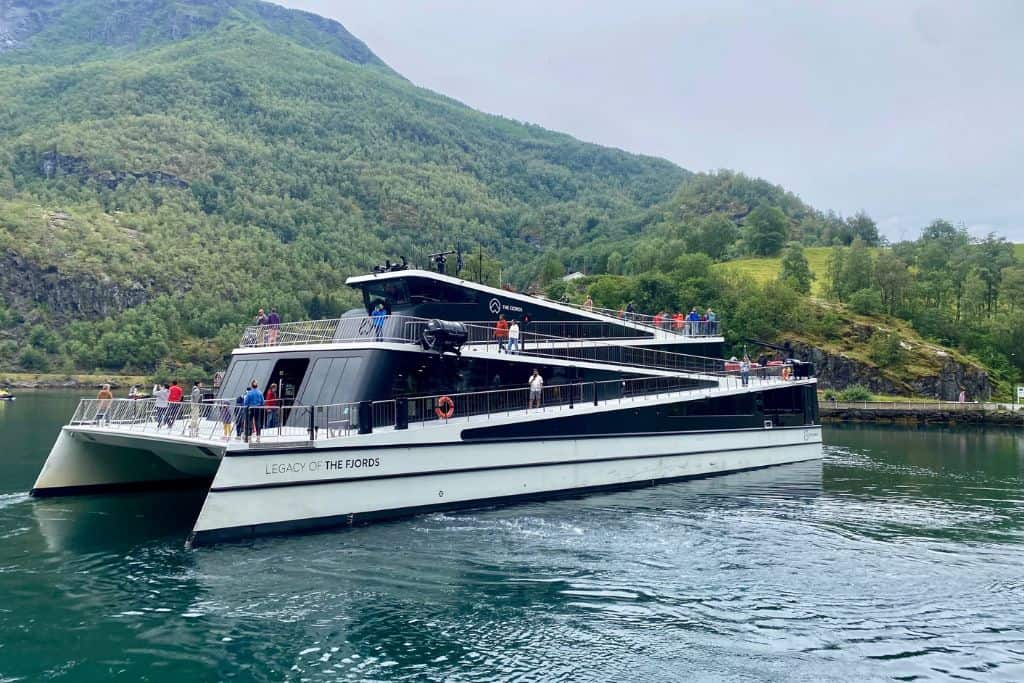 This is an image of an electric fjord cruiseboat that is just leaving Flam to take people into the fjord. In the background are green mountains and on the boat are people who are about to go out on the boat.