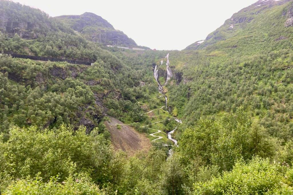 This is a view of green mountains and in the middle of two of them is a track that has 21 hairpin bends. This is in Flam.