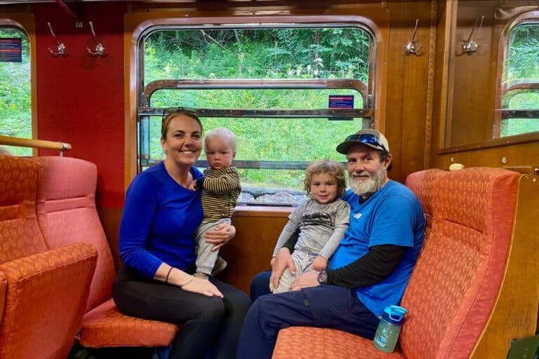 A family are sat in a steam train. On the right is the dad in a blue t-shirt and next to him is his 3 year old son. Opposite them is the mum also in a blue top with their 1 year old son on her lap. Behind them is the train window looking out over the surrounding area. The Flam train is one of the most popular things to do there.