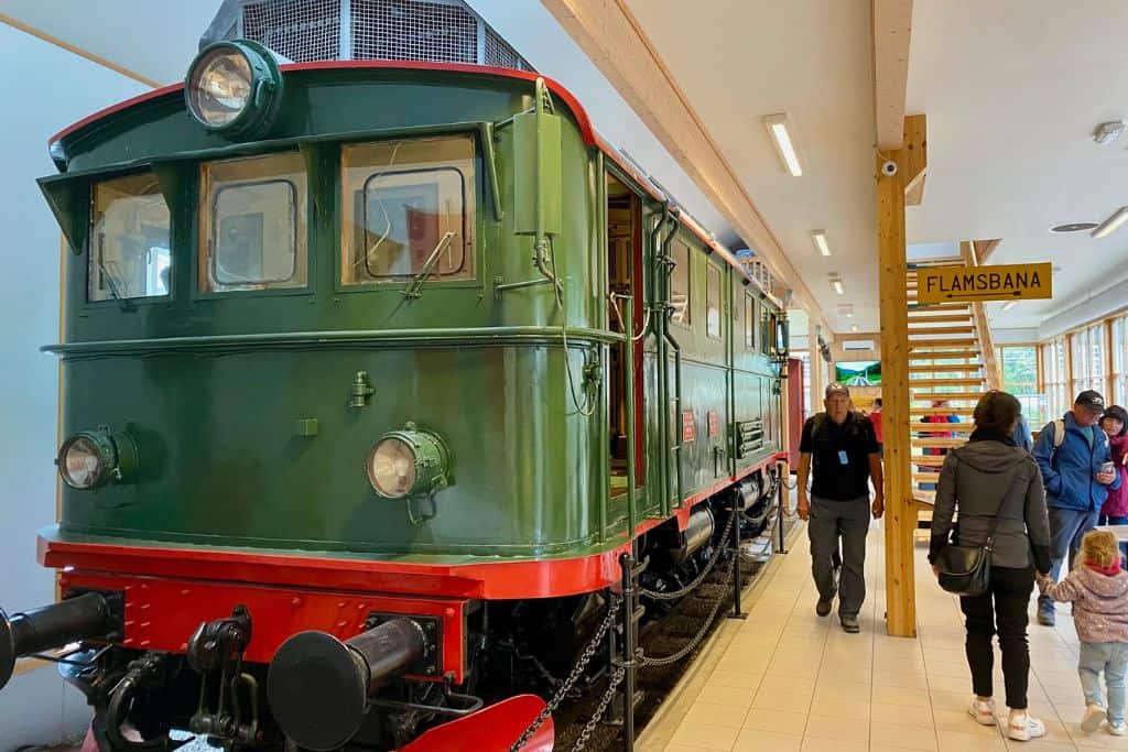 This is a photo of a museum.  There is a green stream train facing the camera with a red underside.  There are people in the museum looking at the train.  The train is one of the trains that used to ride the Flam railway.