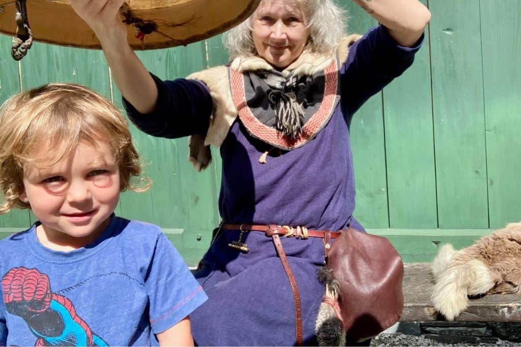 A small boy in a blue spiderman t-shirt is facing the camera.  A woman dressed as a Viking is holding a drum about his head. This is at Viking Valley near Flam which is one of the best things to do with kids.