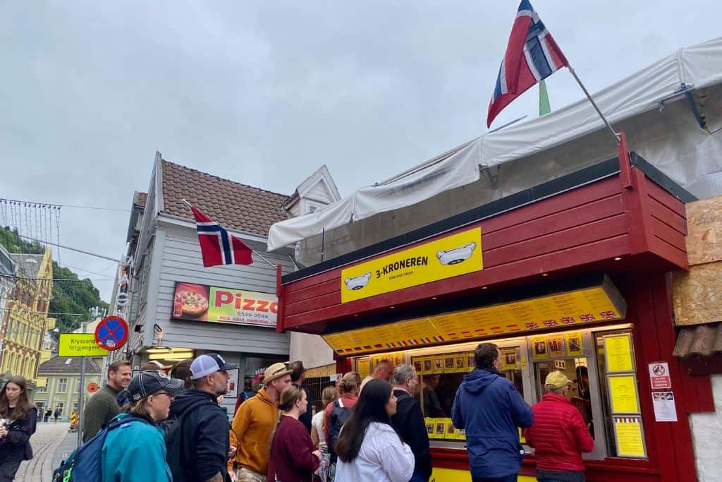 This is the outside of a hot dog stand in Bergen.  There are lots of people queuing up outside of it.