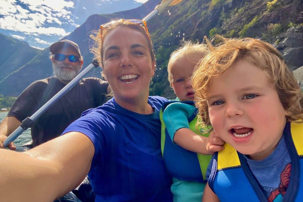 A family are in a kayak and taking a selfie of themselves.  At the back is the dad who is paddling the kayak, in front of his is his girlfriend who is holding the camera and wearing a blue t-shirt.  She is holding their baby who is in a life jacket.  Then at the very front of the image is their 3 year old son who is also in a life jacket and smiling at the camera.  They are in Flam and behind them are the green mountains of the fjord. Kayaking in Flam is one of the best things to do there.