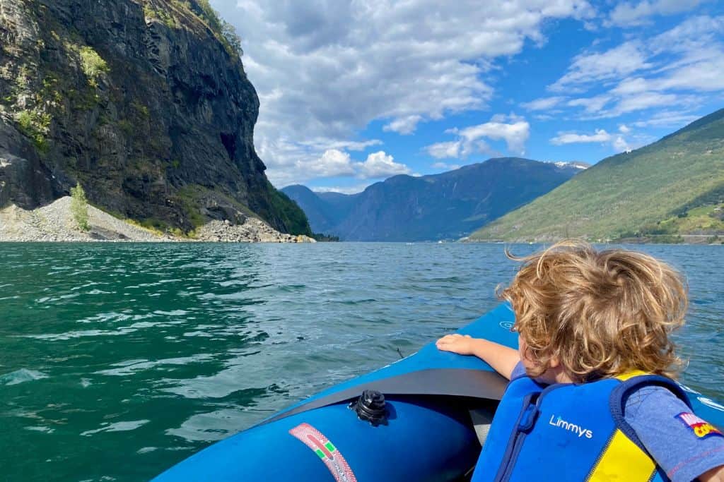 The photo is of the back of a little boy as he looks out over the front of a blue inflatable kayak.  In the background if the fjord.  They are kayaking out from the beach in Flam. 