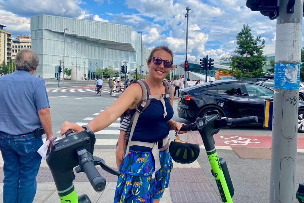 A woman with sunglasses on is smiling at the camera.  On her back is her baby son who is in his carrier asleep. She is holding the handles of an e-scooter in Oslo.