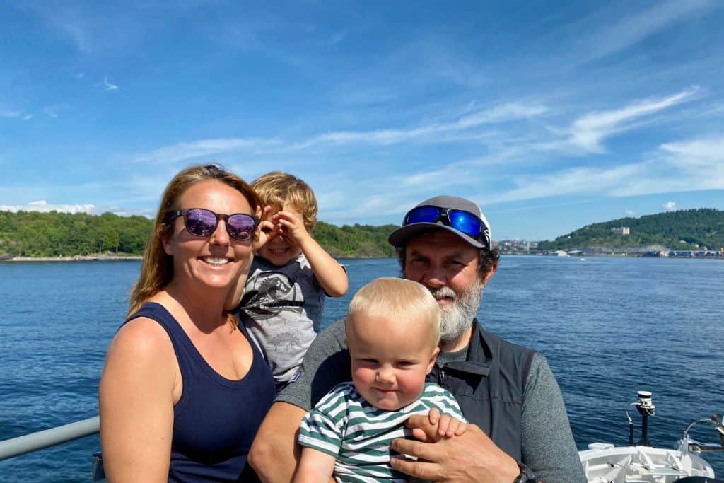 A mum, dad and their two young kids are smiling at the camera.  They are on a ferry boat in Oslo. 