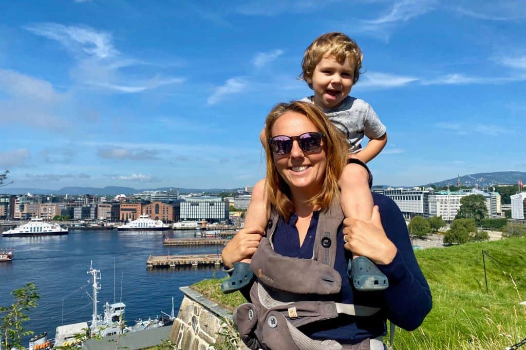 A woman has her sone on her shoulders and they are both looking at the camera.  They are in oslo for one day in the summer and they are on the edge of the fort looking over the waterfront.