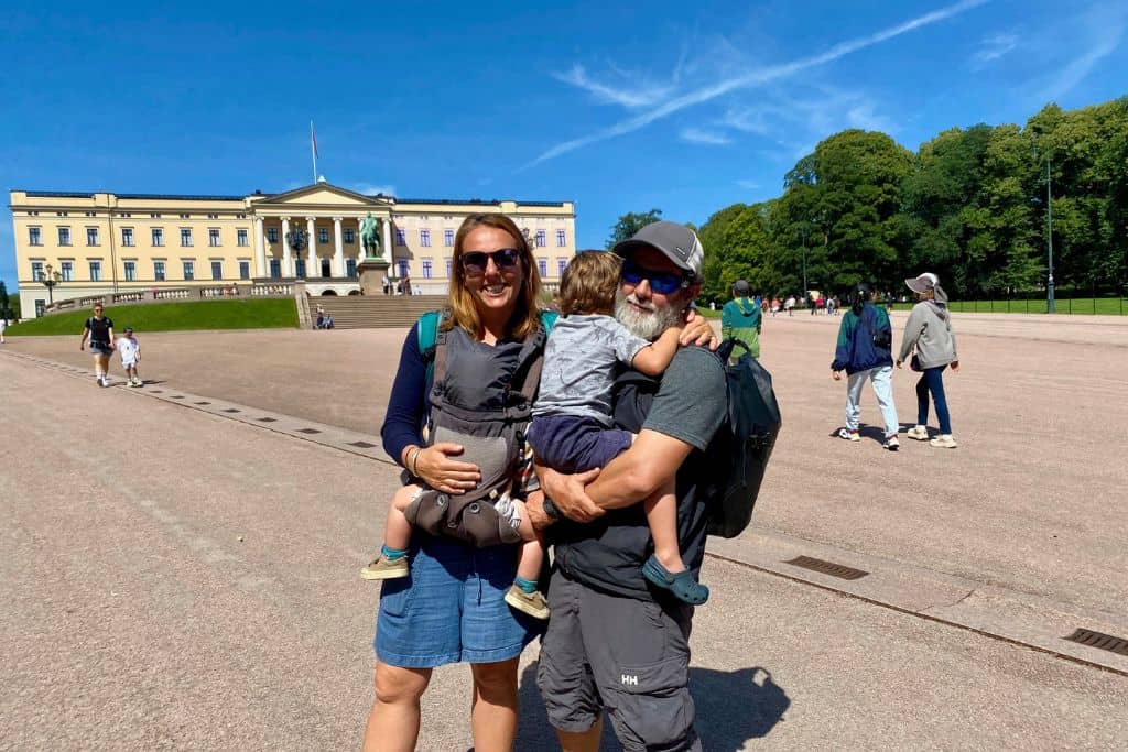 A woman with sunglasses on is smiling at the camera and on her front she has her baby son in a carrier. Next to her is her boyfriend who is holding their other young son who is giving his dad a tight cuddle. They are smiling at the camera and behind them is the Royal Palace in Oslo which they are visiting as part of their one day in Oslo.