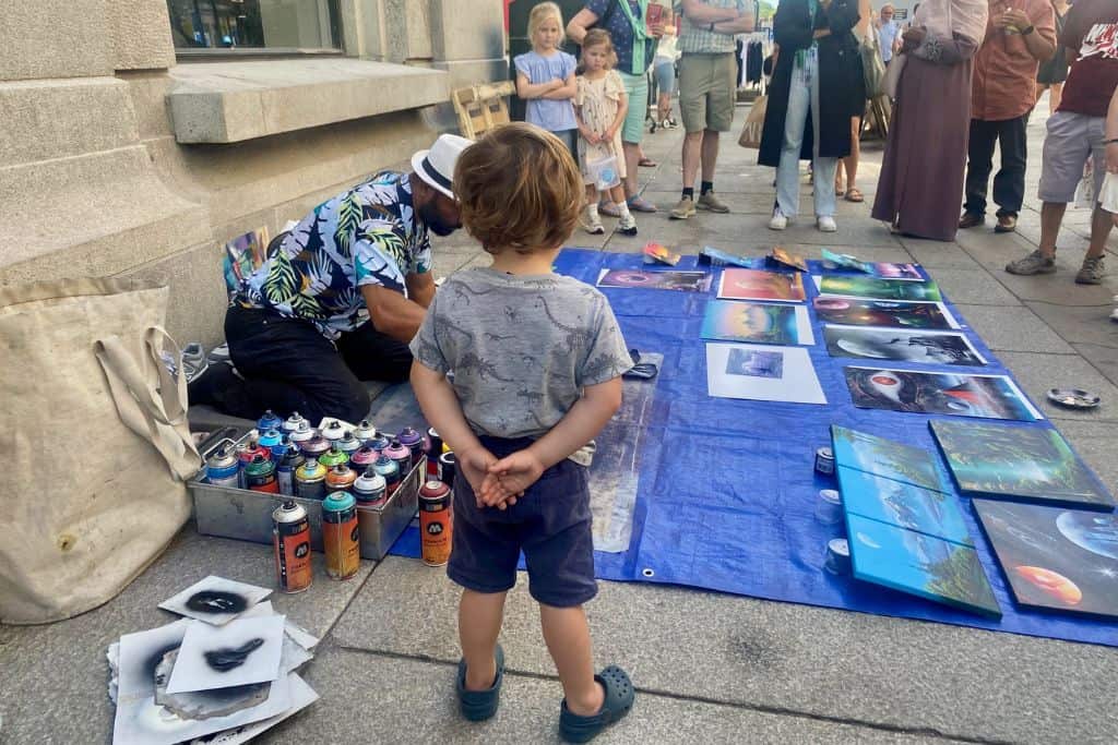 A little boy wearing a grey dinosaur t-shirt with his back to the camera. He is watching an artist spray painting a picture on the ground. This is Karl Johans Gate in Oslo.