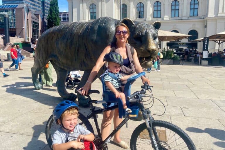 A woman wearing sunglasses, vest top and shorts is smiling at the camera. She is holding the handles of her bike and sat on the front is her baby son wearing a cycle helmet and also looking at the camera. In front of her is her other son who is sat on his bike and looking off into the distance. Behind them all is a huge bronze statue of a tiger. They are cycling past as part of their one day in Oslo.