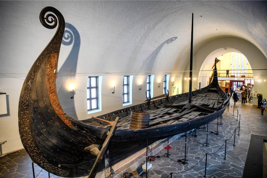 The inside of a museum and in the middle of it there is a huge Viking ship.  This is the Oslo Viking Museum. 