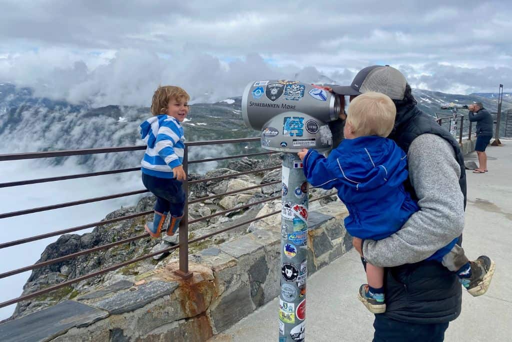 A man is stood holding his baby son in his arms and they are looking through the view finder of a large set of binoculars that you find at tourist attractions.  They are looking into the distance past a wire fence.  Stood on the fence is a 3 year old boy who is looking at the man and baby and laughing.  In the background is a blanket of cloud over Geirangerfjord.  They are at the Geiranger Skywalk which is the best thing to do in Geiranger for panoramic views.