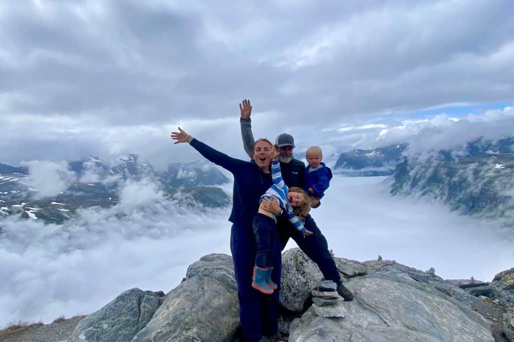 A woman, her boyfriend and their two young kids are stood on top of a rock. They are up so high that behind them is a blanket of cloud as they have gone above the cloud.  They are at the Geiranger skywalk which is one of the best things to do in the village. 