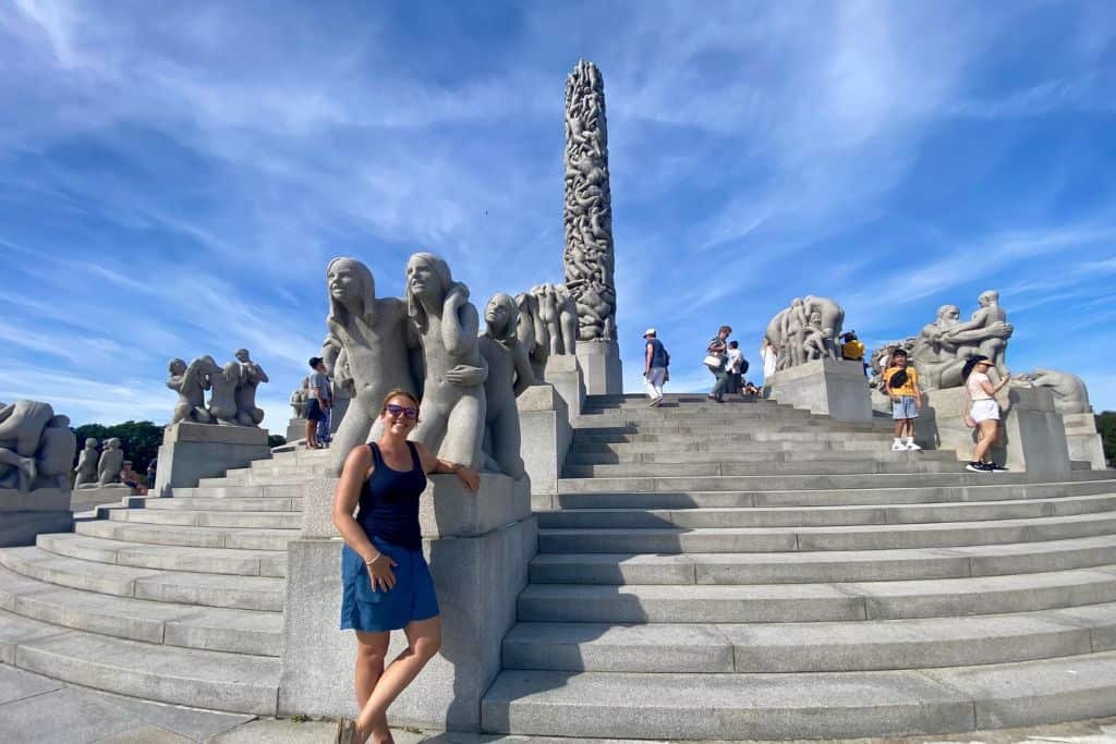 A woman with sunglasses on is smiling at the camera with her arm resting on a statue.  Behind her are lots of statues of people doing different things. In the middle is a totem pole of human figures intertwined. Visiting this park called Vigeland Park is one of the things to do if you have one day in Oslo. 