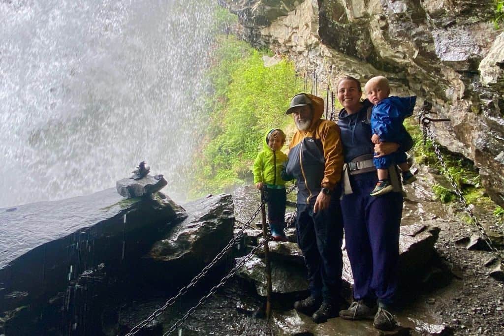 A mum and dad are stood in their raincoats and hiking gear. The mum is holding the baby in a blue waterproof and next to the dad is their three year old boy.  They are all stood on rocks under a waterfall as they are behind the waterfall and it's falling behind them.  This is a waterfall hike in Geiranger and one of the best things to do there.