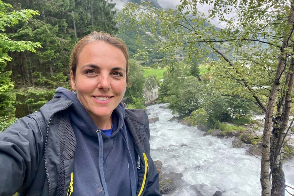 A woman in her raincoat is stood smiling at the camera as she takes a selfie. Behind her are some waterfalls.  She is in Geiranger on the waterfall trail which is an easy hike. 