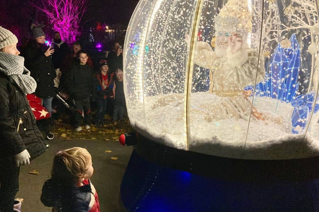 A little boy in the bottom left of the photo is looking up a woman who is dressed up in costume as a snow globe.  It's dark outside and the snow globe is all lit up.