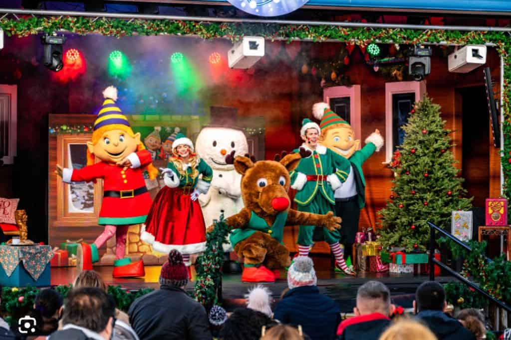 The photo is of a theatre stage made up of actors in costumes, a reindeer, snow man and lots of elves.  The actors are dancing and celebrating the festive season at Paultons Park.  Their Christmas event is one of the best things to do in Hampshire at Christmas.