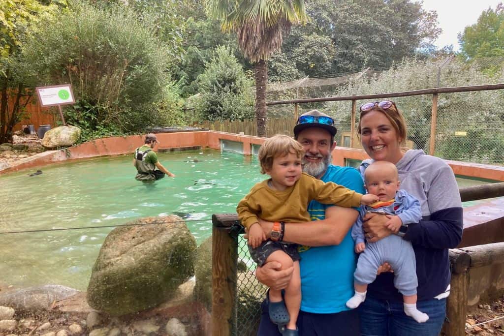 A mum and dad are smiling at the camera and holding their baby and toddler son. They are at the zoo and behind them is a large tank with some penguins in it and a zookeeper is feeding them. 