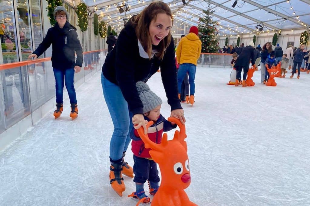 A mum is on an ice rink holding the hands of her very young son who is stood on the ice in front of her using a plastic reindeer to help him skate.  They are ice skating at the Southampton Christmas market which is one of the best things to do in Hampshire at Christmas.