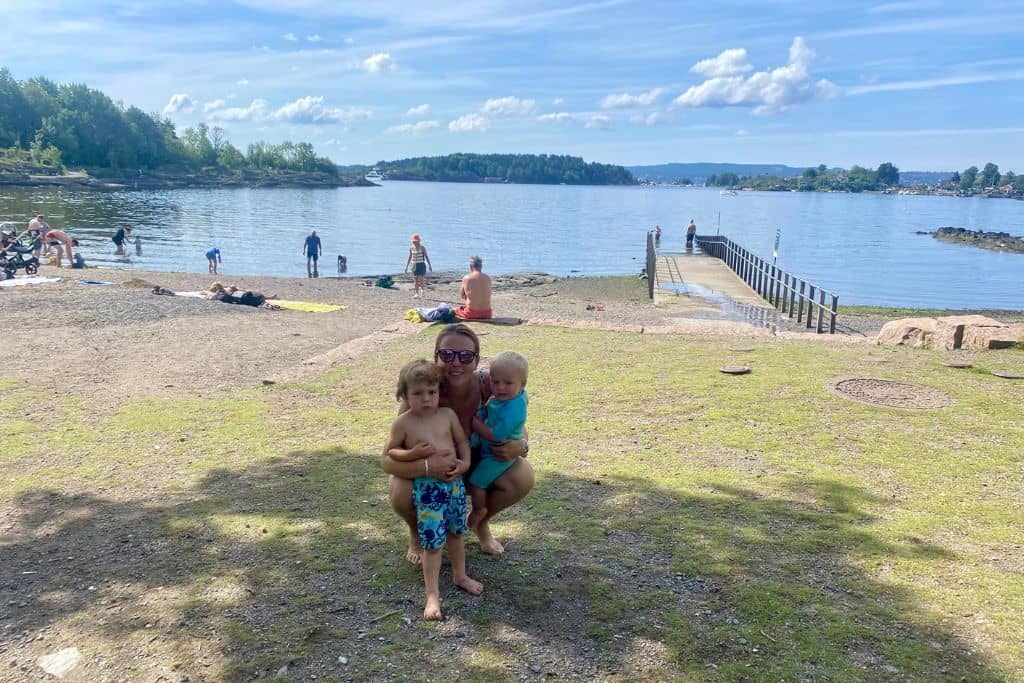 A woman is crouched down on a patch of grass with one of her sons on either side of her. In the background is a stone beach and they are in their swim wear and about to go swimming. There are lots os great small beaches in Olso that you can visit with kids.