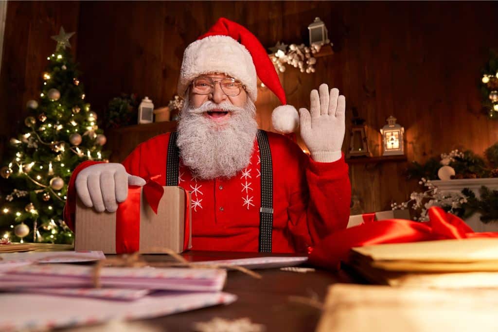 A photo of a man dressed as Father Christmas looking at the camera with his hand up in a high five waving at the camera. On his desk in front of him are some Christmas cards that he's writing. 