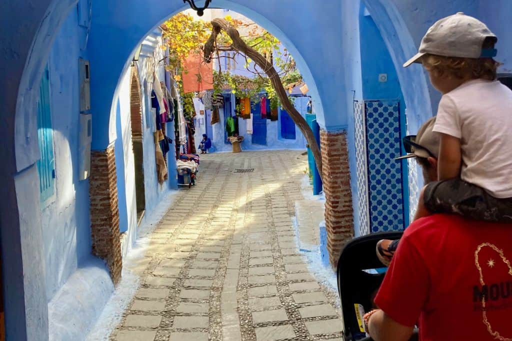 This is a photo of a blue alleyway in the medina in Chefchaouen.  There is an archway and path running through it.  To the right of the image in the foreground is the back of a man with his son on his shoulders as they walk along the streets. 