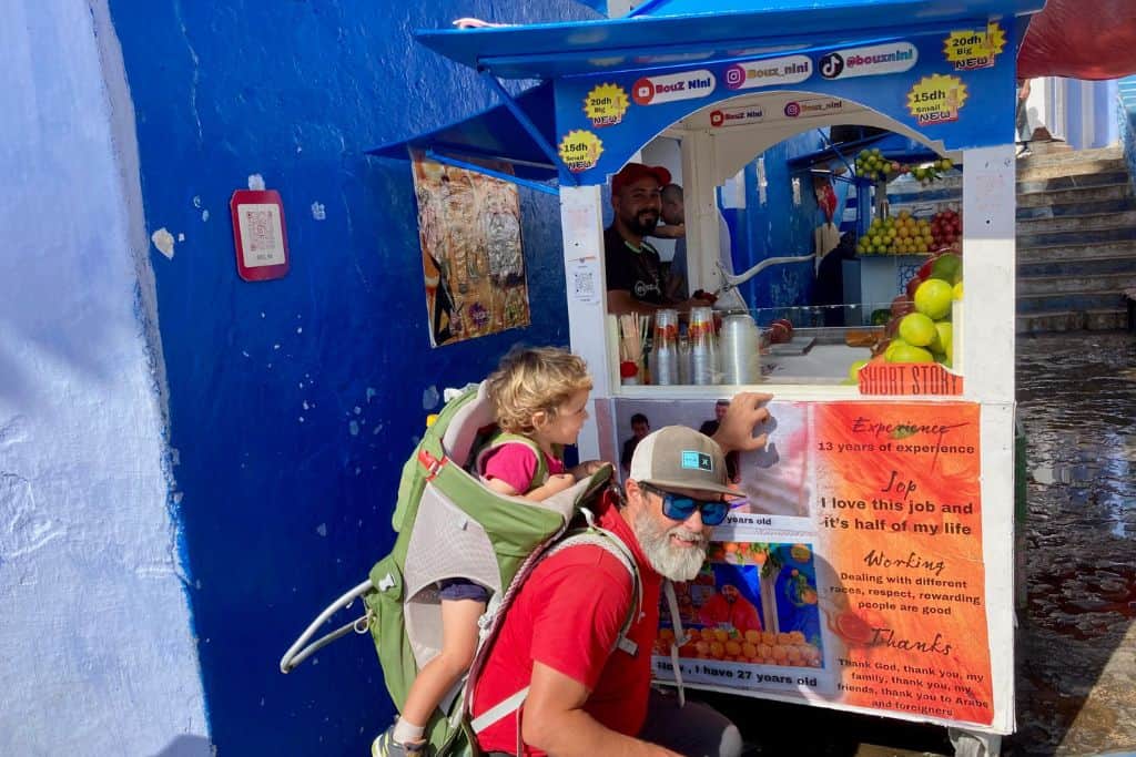 A man in a red t-shirt is crouching down in front of a colourful juice stand in Chefchaouen.  On his back in a green backpack carrier is his son who is also in a red t-shirt.