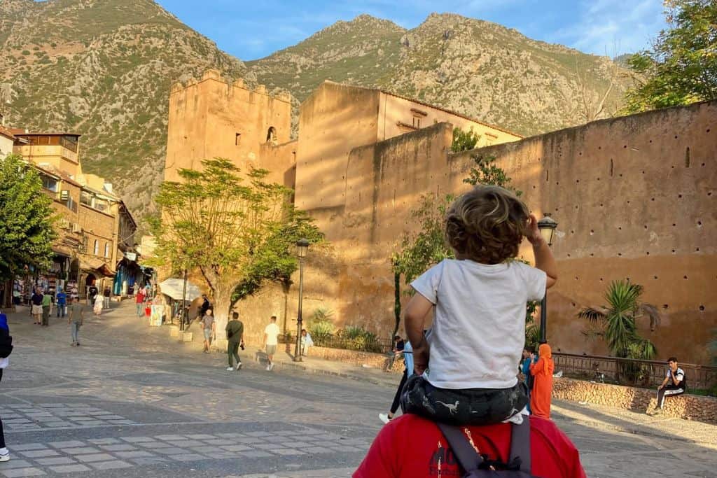 This is a photo of the stone outside of a kasbah. In the foreground a man has his back to the camera and is carrying his son on his shoulders. They are in Chefchaouen which is very family friendly means it is worth visiting with kids.