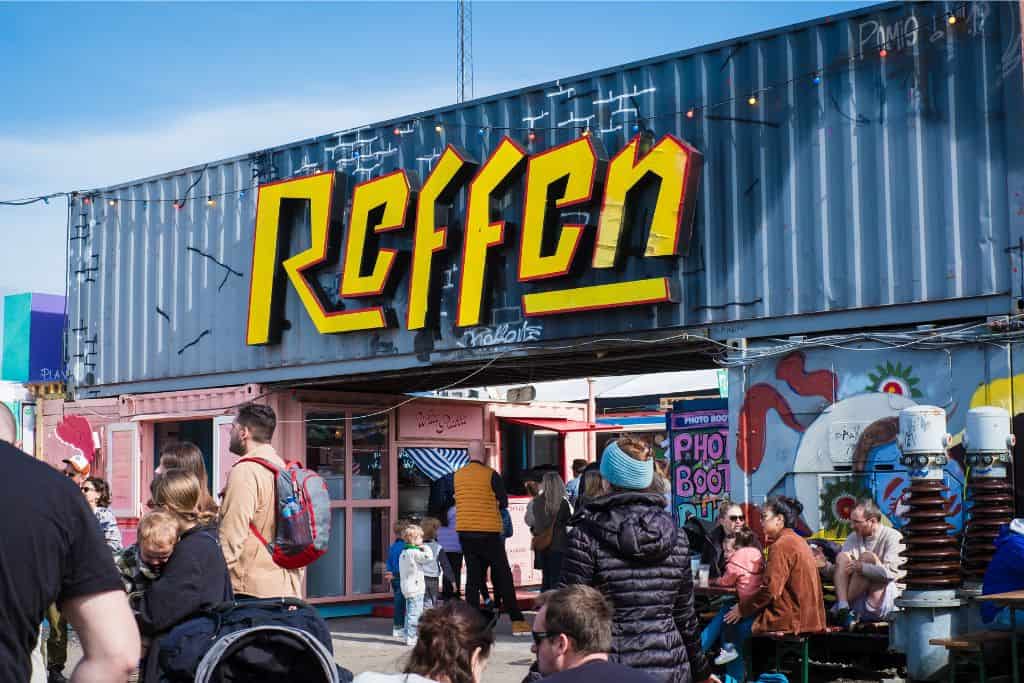 This photo is take from outside Reffen which is a street food market in Copenhagen. There is a yellow sign on a shipping container with Reffen on it.  And below there are people crossing under it and walking through the entrance. Reffen is one of the fun free things to do in Copenhagen.